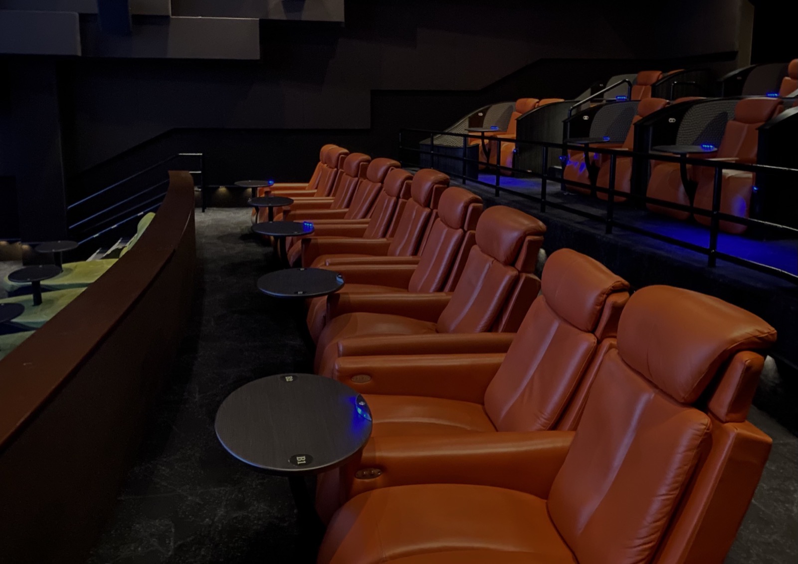 A row of orange leather reclining seats with small side tables in a dark movie theater auditorium