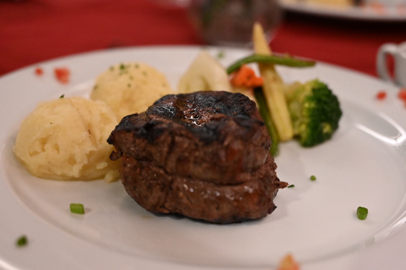 A char-grilled filet mignon on a white plate with mashed potatoes and steamed vegetables