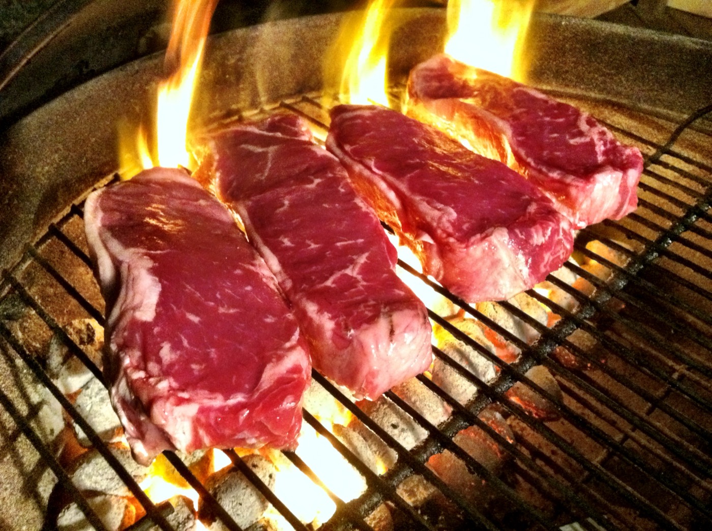 Four raw strip steaks on a grill grate over glowing charcoal with flames rising behind them