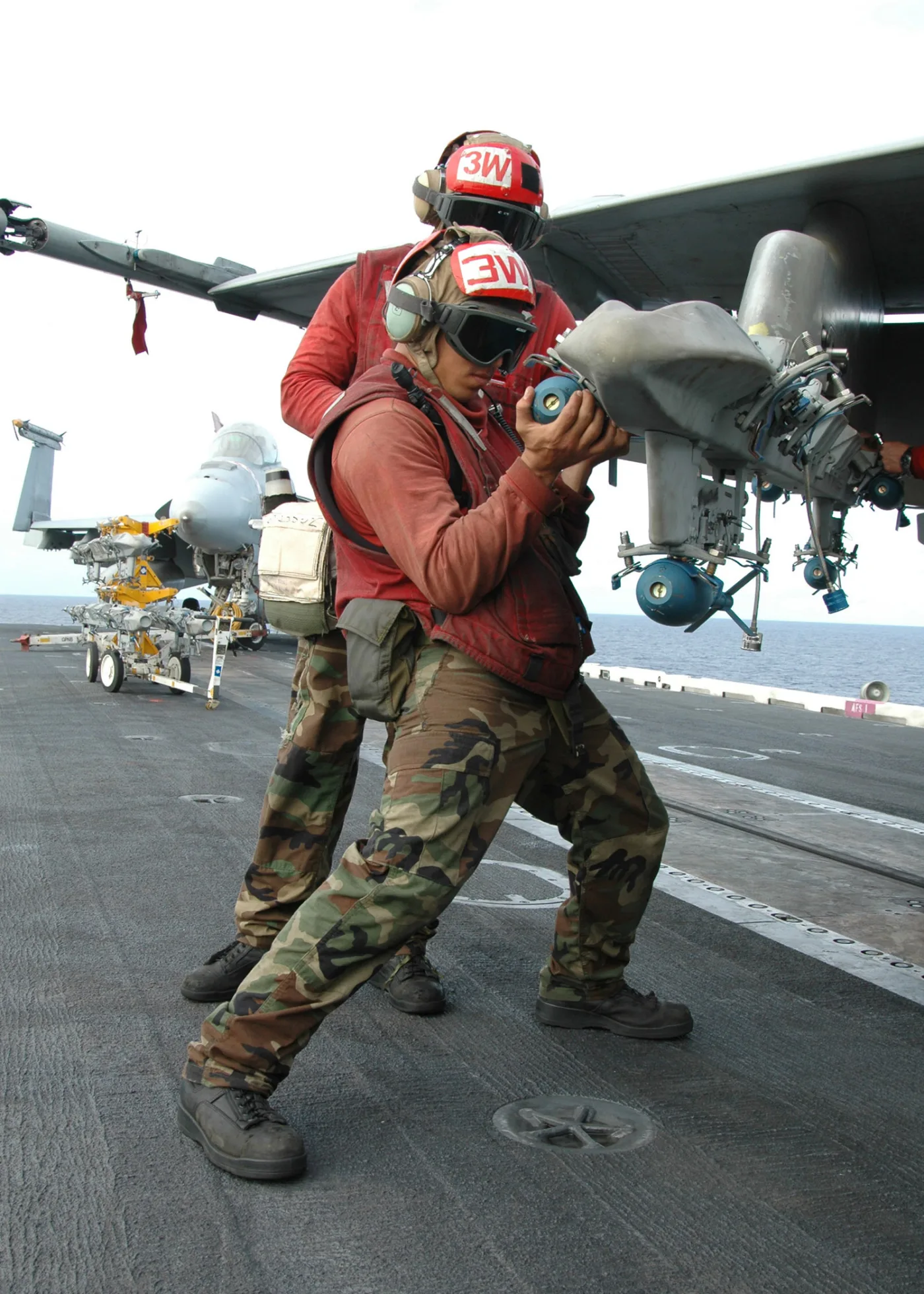 Navy aviation ordnancemen load a small blue MK-76 inert practice bomb onto the wing rack of an F/A-18C Hornet on an aircraft carrier deck